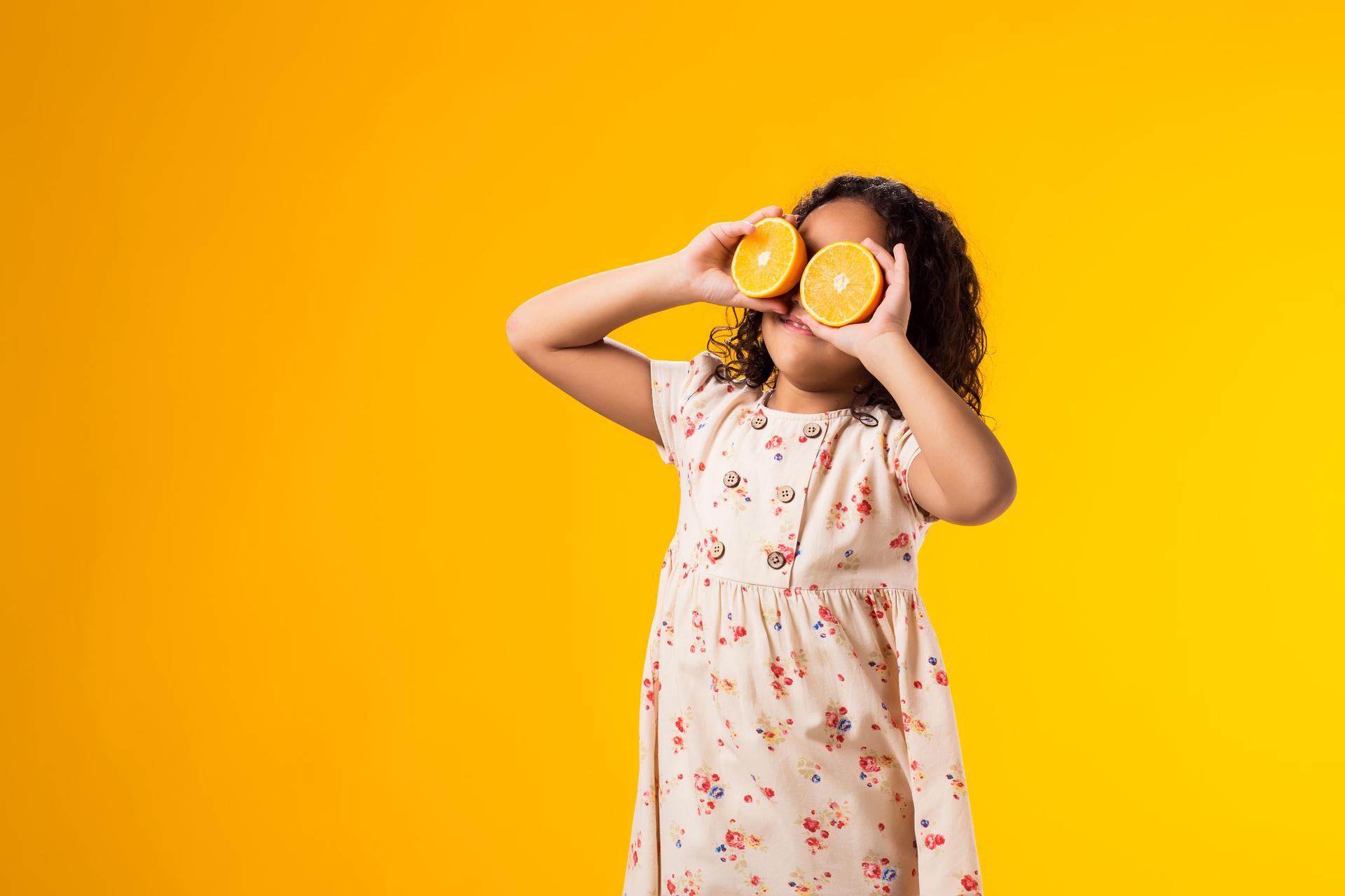 Smiling kid girl holding half of oranges near the eyes. Healthy food, vitamins and children nutrition concept